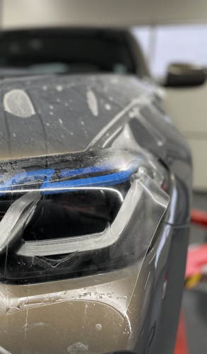 Detailaufnahme bei der Anbringung von Lackschutzfolie im Bereich der Scheinwerfer eines grauen BMW.